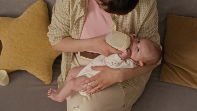 High angle shot of young mother sitting on couch at home and bottle feeding Caucasian infant girl with baby formula