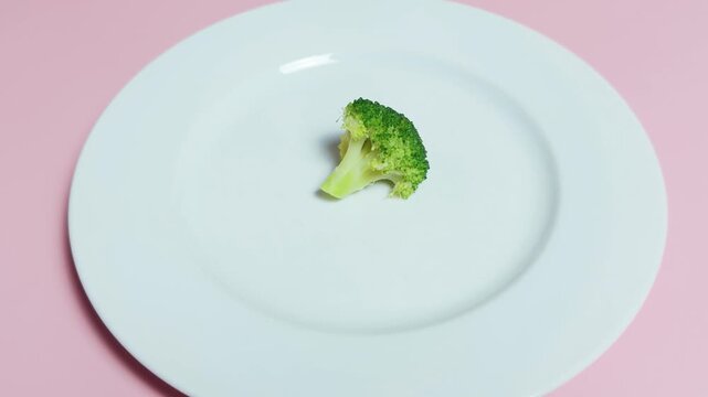 Small green broccoli floret resting on a minimalist white dinner plate against a soft pink background, representing themes of weight loss, restrictive eating, nutrition, and limited food portions