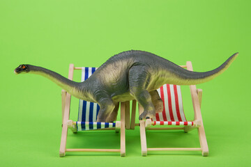 Funny toy sauropod dinosaur trying to fit on two miniature beach deck chairs, green background. © aneduard