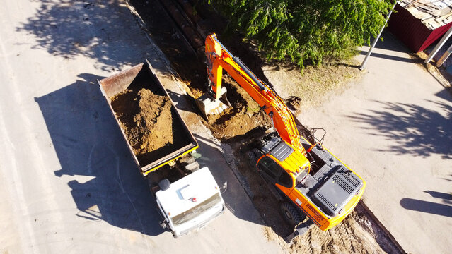 A high-angle drone shot capturing an orange hydraulic crawler excavator digging a trench and loading soil into the back of a white tipper dump truck