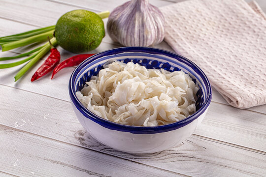 Fresh ingredients for Asian cuisine with a bowl of wide rice noodles, garlic, lime, and chili peppers on a white wooden table.