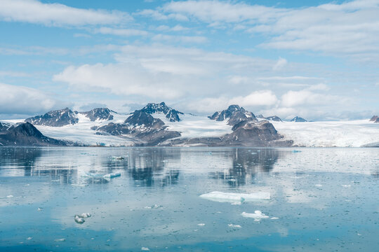 View of Lilliehookbreen glacier with icebergs floating in the calm blue water and rugged snow-capped mountains under a cloudy sky in Svalbard, Svalbard and Jan Mayen.