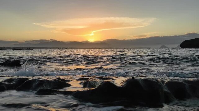 Majestic sunset over the ocean with waves crashing on rocky shore. Golden sun glowing behind clouds with silhouette of mountains and boats, includes ambient wave sounds. Jember Indonesia.