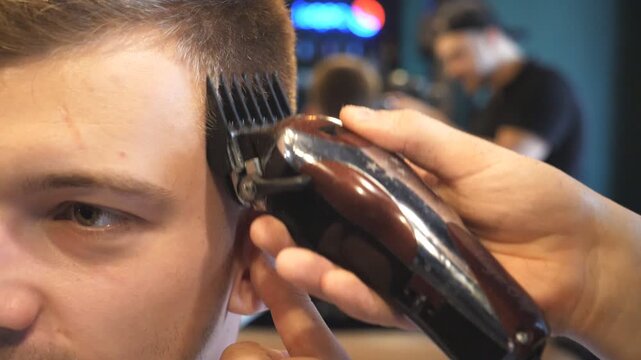 Close up hands of hairstylist cutting hair of young client with electric razor in barbershop. Arms of barber making male haircut to customer with clipper in salon. Barber shaving man with trimmer