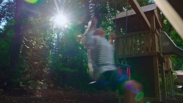 Child swinging on outdoor playset with bright sun flare through forest trees creating nostalgic summer atmosphere focused on freedom imagination and joyful youth