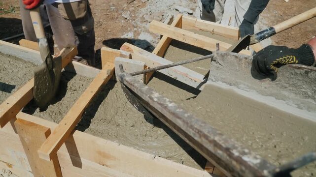 Builders pouring and leveling wet cement in wooden formwork for a house foundation, construction site work.