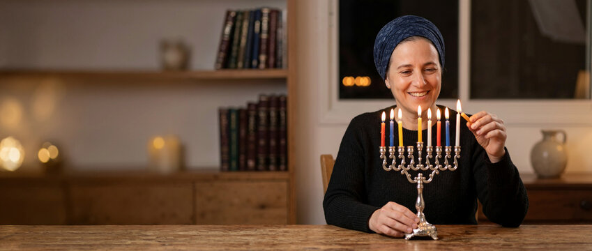 Smiling Jewish woman lighting candles on a silver Hanukkah menorah. Happy female celebrating the festival of lights at home