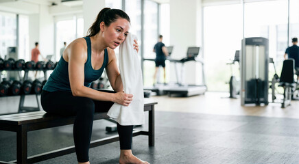 Fototapeta premium Sweaty woman wiping her face with a towel in the gym. Tired female athlete resting on a bench after an intense workout. Fitness and weight loss recovery