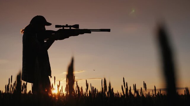 Silhouette of woman holding sniper rifle with scope at sunset in wheat field.