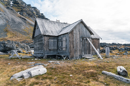 View of the historic wooden cabin at Varsolbukta surrounded by rocky tundra and driftwood under a cloudy sky with mountains in the background Svalbard, Svalbard and Jan Mayen.