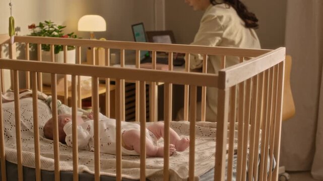 Side view slowmo shot of young mother working on laptop from home in evening, sitting in cozy nursery room near infant baby girl lying in wooden crib in foreground