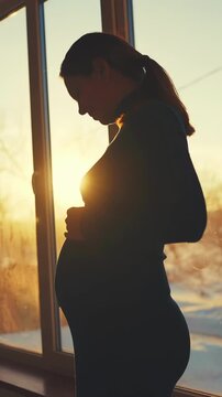 A woman stands near a window, silhouetted against the sunset, holding her pregnant belly. The warm sunlight casts a glow around the woman and highlights her pregnancy.