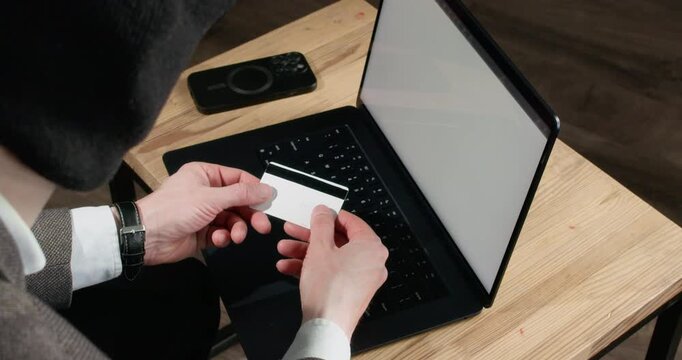 Internet scammer rotates bank card in hands while sitting at laptop during fraud. Man examines card details as preparing to use payment data for deception