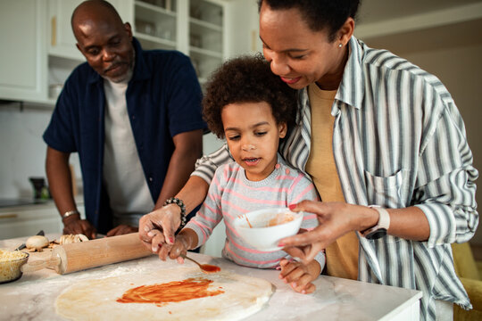 Parents help child make pizza in a home kitchen