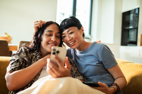 Two women relaxing on a sofa at home, smiling at a smartphone