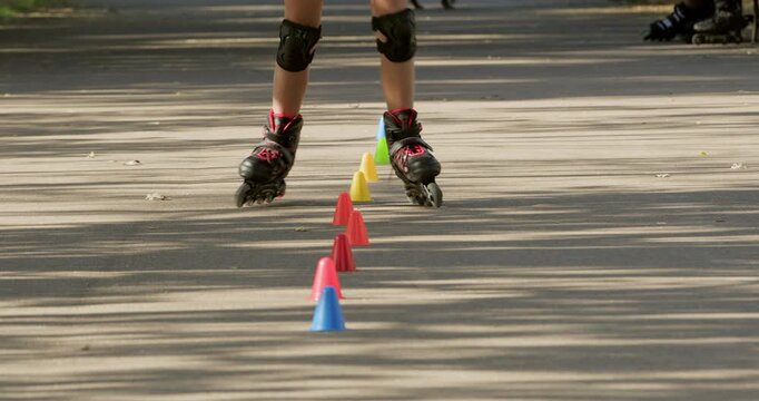 Girl rides inline skates passing colorful cones on park road during training. Kid in pads keeps feet aligned sliding between markers with controlled rollerblade moves