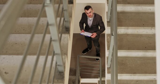 Architect studies blueprints and gives recommendations to workers. Businessman in tailored jacket carefully walks down concrete staircase and reviews technical papers