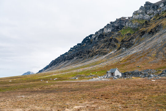 View of a small wooden cabin at the base of a steep rocky mountain in the tundra landscape of Varsolbukta Svalbard, Svalbard and Jan Mayen.