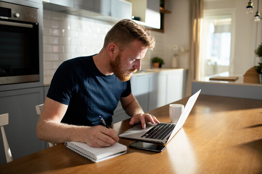Man working on laptop and taking notes at kitchen table