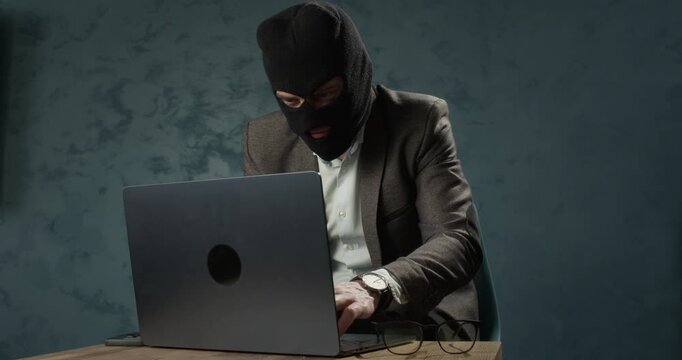 Masked man sits at laptop while typing during online scam operation. Fraudster uses computer as hiding identity behind balaclava and dark clothing in room