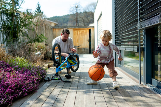 Toddler playing basketball while father repairing bicycle at home terrace.