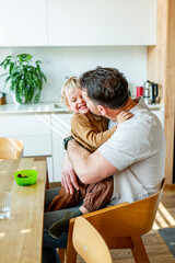 Dad feeding toddler during family mealtime in modern kitchen.