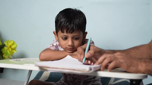 Young Indian boy looking bored while studying at a desk with a teacher's hand nearby, Delhi, India