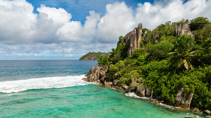 Obraz premium Turquoise waters meeting rocky cliffs surrounded by dense, lush greenery under a bright sky. Seychelles, Mahe.