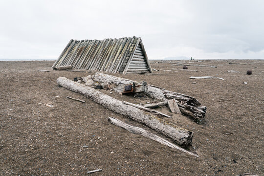 View of the historic A-frame log hut at Poolepynten with scattered driftwood on a desolate gravel beach under an overcast sky in Svalbard, Svalbard and Jan Mayen.