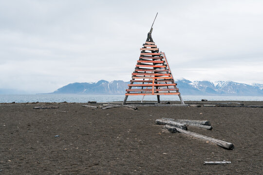 View of the Poolepynten wooden navigation beacon on a dark pebble beach with driftwood and snow-capped mountains in Svalbard, Svalbard and Jan Mayen.