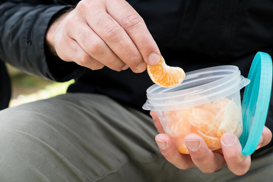 A person holding a plastic food container with a turquoise lid, carefully picking up a single mandarin orange segment, highlighting microplastic contamination concepts.