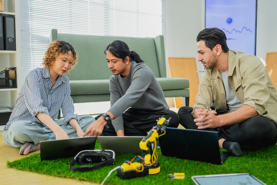 Diverse team of young tech engineers collaborating on robotics development and programming a yellow robotic arm, using laptops while sitting together in a modern startup office workspace, focusing on 