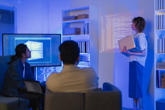 A professional East Asian female software engineer stands holding a laptop and gesturing toward lines of code projected on a wall display, presenting data scripts to her multiethnic colleagues during 
