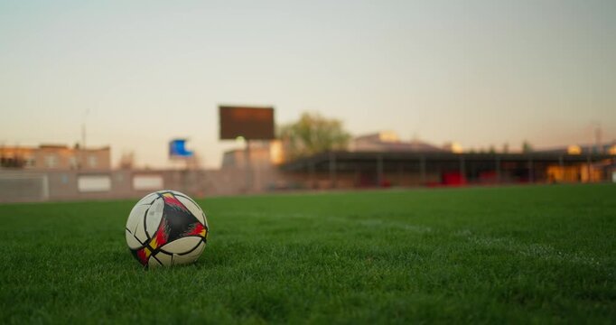 Close-up soccer ball resting on lush grass field at sunset with player preparing to kick, blurred stadium stands and scoreboard in background