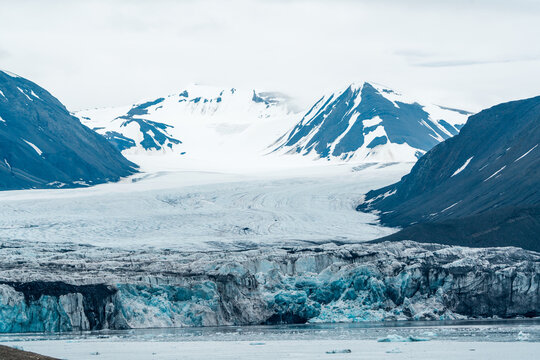 View of a massive blue glacier flowing between snow-capped mountains into the arctic sea under an overcast sky Svalbard and Jan Mayen.