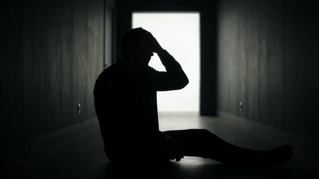 Silhouette of a man sitting alone in a dim hallway, head in hand, representing depression, isolation, sadness, mental struggle, solitude, stress, and heavy burden