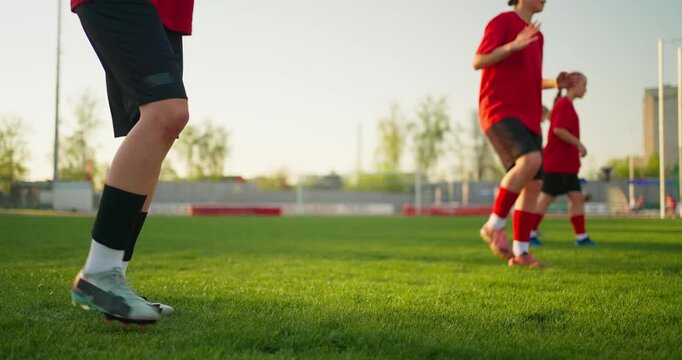 Youth soccer players in red jerseys warming up on a sunlit grass field during team practice, with focus on legs, cleats, and training drills