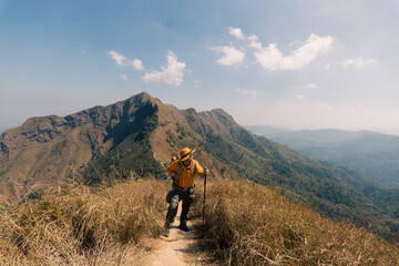 Obraz premium Hiker with yellow backpack and hat standing on a grassy hill, looking at a majestic mountain ridge. Adventure, trekking, and exploration in the wild nature concept.