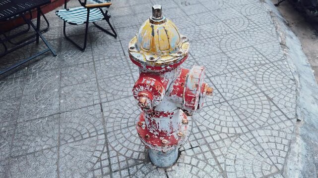 Worn red and yellow fire hydrant stands on a patterned sidewalk in an urban setting, representing city infrastructure, emergency services, and the visible effects of time on public utilities