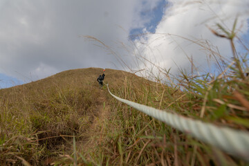 Obraz premium A long-distance view of a lone hiker walking along a narrow, grassy mountain ridge path, guided by a prominent safety rope in the foreground, with layers of hills fading into the background.