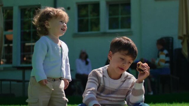 Toddler standing beside older sibling reading illustrated book on sunny grass lawn while holding burger conveying playful childhood bonding family leisure and outdoor relaxation