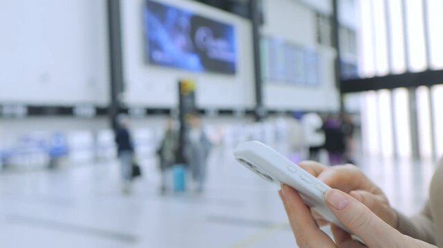 Hand holds smartphone at busy airport terminal. Blurry travelers move in background. Large screen displays flight information. Bright natural light from windows. Modern architecture with high ceilings