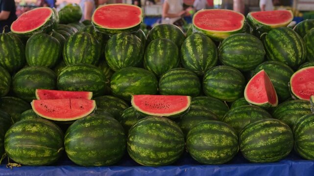Row of stacked watermelons at market, ripe red slices displayed on blue tablecloth, striped green rinds and juicy flesh, vendor arranging produce, blurred