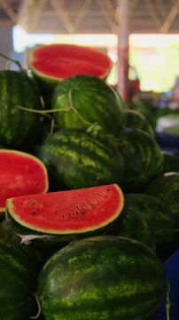Row of stacked watermelons at market, ripe red slices displayed on blue tablecloth, striped green rinds and juicy flesh, vendor arranging produce, vertical