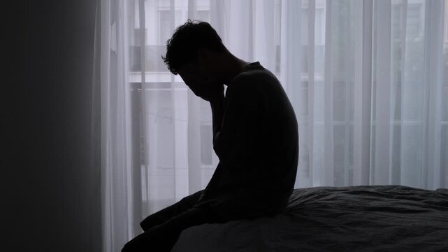 Burnout and emotional exhaustion silhouette of lonely young man sitting on bed near window feeling depressed disconnected and mentally overwhelmed at home