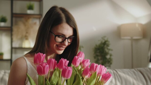Young woman with glasses smiles while smelling a bouquet of vibrant pink tulips indoors