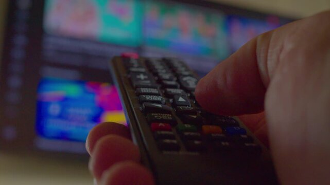 A man's hand uses a remote control to change the content on a modern wall-mounted television. First-person view. Close-up. Shallow depth of field. Video shot in natural light.