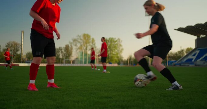 Female soccer players in red uniforms practice drills on a green stadium field at sunset while a referee jogs beside the action