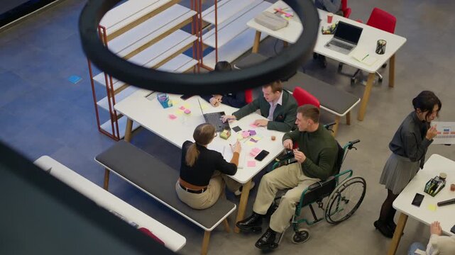A diverse business team actively collaborates on a project during an office meeting. Colleagues, including one in a wheelchair, are working together in a modern workplace.