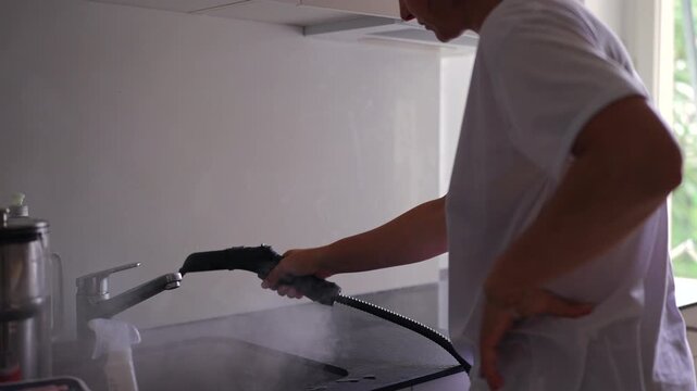 Woman steam cleaning modern kitchen sink and countertop capturing hygiene, deep sanitation and authentic everyday household maintenance routine indoors